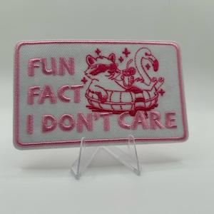 Op de afbeelding: Een rechthoekige geborduurde patch met een witte achtergrond en roze rand. Het toont een wasbeer met een zonnebril in een flamingo-zwemband, die een drankje vasthoudt. De tekst "FUN FACT I DON'T CARE" is in roze letters geborduurd.