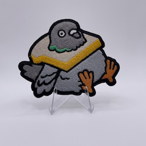 Op de afbeelding: Geborduurde patch met een cartoon duif die een sandwich draagt. De duif is grijs met oranje voeten en een zwarte omtrek. De sandwich is bruin met een gele korst en groene vulling. De patch wordt gepresenteerd op een transparante plastic standaard.