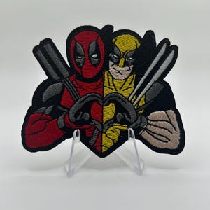 Könnte beinhalten: Ein gestickter Aufnäher mit einem geteilten Design von Deadpool in Rot und Schwarz auf der linken Seite und Wolverine in Gelb und Schwarz auf der rechten Seite. Beide Figuren formen mit ihren Händen ein Herz, mit Schwertern im Hintergrund.