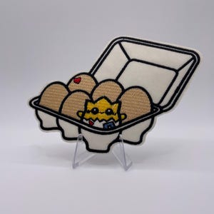 Può includere: Toppa ricamata raffigurante un cartone di uova marroni, con un pulcino giallo che esce da una di esse. Il cartone delle uova è bianco con un contorno nero. Un uovo ha un cuore rosso. Il pulcino indossa un bavaglino blu.