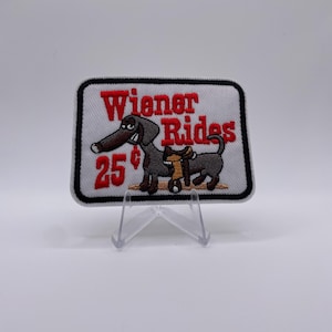 Op de afbeelding: Geborduurde patch met de woorden "Wiener Rides" in het rood, met een cartoon teckel met een zadel. De patch toont ook de prijs "25¢". De patch heeft een witte achtergrond met een zwarte rand.