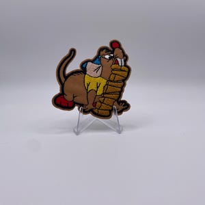 Op de afbeelding: Geborduurde patch met een cartoon bever in bruin, geel en blauw, die een stapel boomstammen vasthoudt. De patch heeft rode accenten op de voeten en het hoofd, met een zwarte rand. De patch wordt gepresenteerd op een transparante standaard.