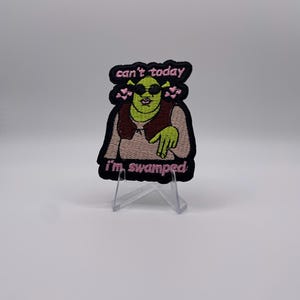Op de afbeelding: Geborduurde patch met een groene oger met zonnebril en vest, met de roze tekst "can't today, I'm swamped". De patch heeft een zwarte rand en wordt gepresenteerd op een transparante standaard.
