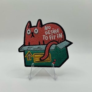 Könnte beinhalten: Ein bestickter Aufnäher mit einer roten Katze mit großen Augen, die in einem grünen Pappkarton sitzt. Der Text NO DESIRE TO FIT IN ist in weißen Buchstaben über der Katze gestickt. Der Karton hat ein gelbes Etikett und ein gelbes Pfeilsymbol.