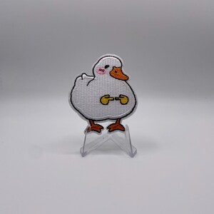 Op de afbeelding: Een witte eendenpatch met een zwarte omtrek, oranje snavel en poten en roze wangen. De eend houdt twee gele voorwerpen vast. De patch wordt weergegeven op een doorzichtige plastic standaard.