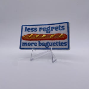 Op de afbeelding: Geborduurde patch met de tekst "less regrets more baguettes" in blauwe letters. De patch heeft een illustratie van een baguette in bruin- en geeltinten. De patch heeft een witte achtergrond met een blauwe rand.