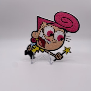 Op de afbeelding: Geborduurde patch met een stripfiguur met roze haar en grote roze ogen. Het personage lacht en houdt een gouden sterrenvormige toverstaf vast. De patch heeft een witte rand en wordt gepresenteerd op een transparante standaard.