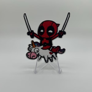 Può includere: Una toppa ricamata raffigurante un personaggio rosso e nero con occhi bianchi che impugna due spade argentate mentre cavalca un unicorno bianco con muso rosa, criniera multicolore e un piccolo cuore rosso sul fianco, il tutto bordato di nero.