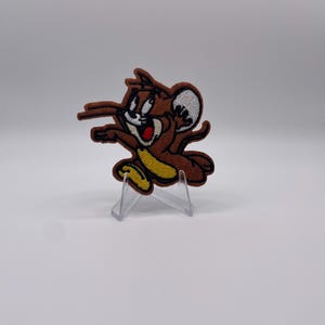 Könnte beinhalten: Gestickter Aufnäher mit Jerry Maus aus Tom und Jerry. Der Aufnäher zeigt Jerry in einer Laufpose, mit braunem Fell, einem weißen Ohr und einem gelben Bauch. Der Aufnäher wird auf einem transparenten Ständer vor weißem Hintergrund präsentiert.
