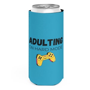 May include: A blue can cooler with the words "ADULTING ON HARD MODE" in black text. A yellow video game controller is below the text. The cooler is designed to hold a standard-sized beverage can.
