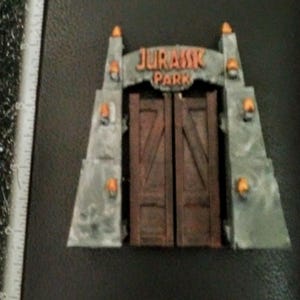 May include: A miniature model of the Jurassic Park gate, featuring a stone archway with the words "Jurassic Park" in orange letters and two brown wooden doors.