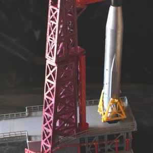 May include: A red metal model rocket launch tower with a silver model rocket attached to the top. The rocket is on a yellow metal base.