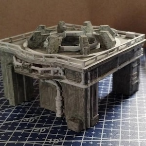 May include: A miniature architectural model of a stone structure with a square base and a circular central feature. The model has a weathered, gray-green color with white accents, and a detailed railing around the top.