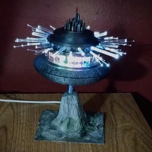 May include: A 3D printed model of a futuristic city, with a black base and a silver dome. The city is lit up with white fiber optic lights, creating a glowing effect. The model is mounted on a wooden base.