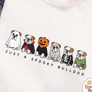 以下が含まれることがあります： 幽霊、吸血鬼、カボチャ、ミイラ、骸骨、モンスターのハロウィーンの衣装を着た6匹のイングリッシュ・ブルドッグのグラフィックが描かれた白いTシャツ。犬の下には「JUST A SPOOKY BULLDOG」というテキストがプリントされています。