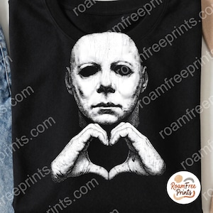 May include: A black t-shirt featuring a white graphic print of a pale, textured mask with dark, hollow eyes. The figure is positioned with both hands raised towards the chin, fingers touching to form a heart shape. The design is centred on the chest.