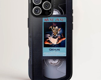 May include: Black phone case featuring a VHS tape design with the text "WE'RE HERE!" and a movie poster for "Gremlins." The case is designed for iPhone, Samsung Galaxy S, and Google Pixel phones.