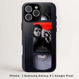 Op de afbeelding: Zwarte telefoonhoes met een VHS-tape-ontwerp, met de filmposter voor "The Lost Boys". De hoes heeft een zwarte rand en de tekst "Insert this side into recorder". Compatibel met iPhone, Samsung Galaxy S en Google Pixel.