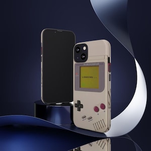 May include: A beige phone case designed to resemble a retro handheld gaming device, with a screen displaying "LOADING...". The case features a D-pad, buttons, and speaker vents. A black smartphone is next to the case.
