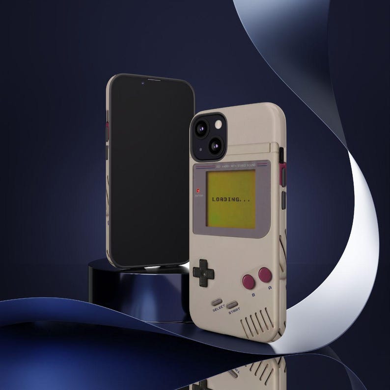 May include: A beige phone case designed to resemble a retro handheld gaming device, with a screen displaying "LOADING...". The case features a D-pad, buttons, and speaker vents. A black smartphone is next to the case.