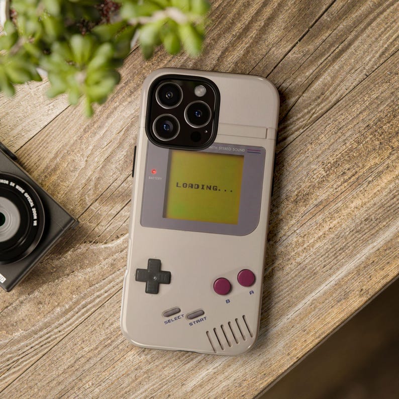 May include: A light gray phone case designed to resemble a classic handheld gaming device. The case features a screen displaying "LOADING...", a directional pad, and pink buttons. The phone case is on a wooden surface.