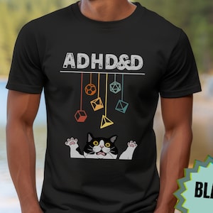 May include: Black t-shirt featuring the text "ADHD&D" in white with a graphic of a black and white cat with yellow eyes and a string of colourful dice hanging above the cat.