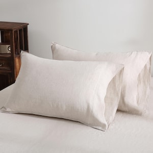 May include: Two light beige linen pillowcases. The rectangular pillowcases have a soft, textured appearance. They are placed on a bed with matching sheets, creating a cohesive, neutral-toned bedding set. The pillows are made of a natural fabric.