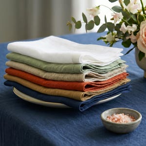 May include: A pile of folded linen napkins in shades of white, green, orange, and blue. The napkins are stacked on a white plate, with a small bowl of pink salt in front. A vase of flowers is visible in the background.