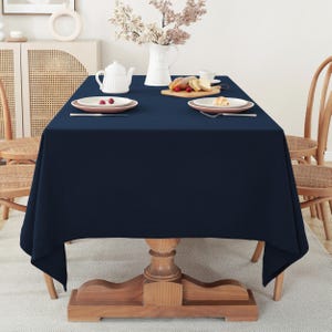 May include: A dark blue rectangular tablecloth is laid on a wooden table with a light wood base. Two white plates with red berries are on the table, along with a white teapot and a plate of food.