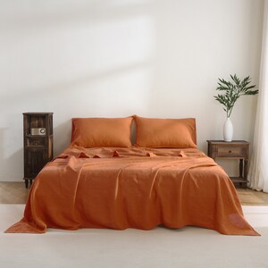 Puede incluir: Un juego de cama de lino de color naranja quemado con dos fundas de almohada y una s&aacute;bana plana. La ropa de cama est&aacute; colocada en una cama con una cabecera blanca y una mesita de noche de madera.