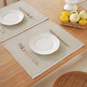 Puede incluir: Dos manteles individuales de lino beige con platos blancos y tenedores plateados sobre una mesa de madera. Un cuenco de fruta se encuentra sobre la mesa.