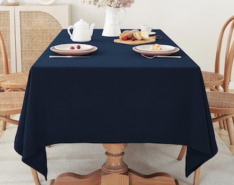 May include: A dark blue rectangular tablecloth is laid on a wooden table with a light wood base. Two white plates with red berries are on the table, along with a white teapot and a plate of food.