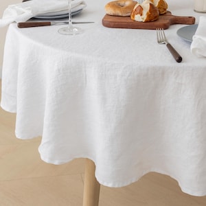 May include: A white, round linen tablecloth draped over a wooden table. The tablecloth has a textured appearance and is set with a plate, silverware, and a wine glass. A wooden cutting board with bread is also on the table.