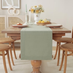 May include: A light green linen table runner with a subtle texture and a white border. The runner is laid on a wooden table with two chairs and a vase of flowers.