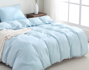 May include: A light blue linen duvet cover with a white throw blanket on top. The duvet cover is on a bed with a white headboard and a white nightstand with a vase of green plants.