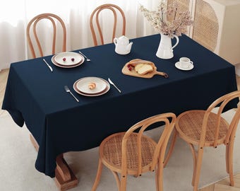 May include: A navy blue tablecloth is laid out on a wooden table with four chairs. The table is set with two white plates, two sets of silverware, a white teapot, a white pitcher, a wooden cutting board with bread, and a white saucer with a cup.