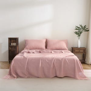 May include: A blush pink linen bedding set with two pillowcases and a flat sheet. The bedding is laid out on a bed with a wooden nightstand and a vase of greenery in the background.