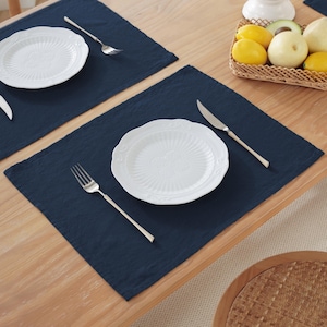May include: A dining table set with two navy blue placemats, each with a white plate, fork, and knife. The plates feature an embossed design. A basket of fruit is visible in the background.