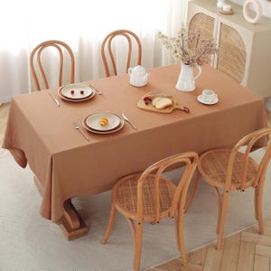 May include: A brown linen tablecloth with a white ceramic teapot, two white plates, a small white cup, and a wooden cutting board with bread on it. The tablecloth is on a wooden table with four wooden chairs with woven seats.