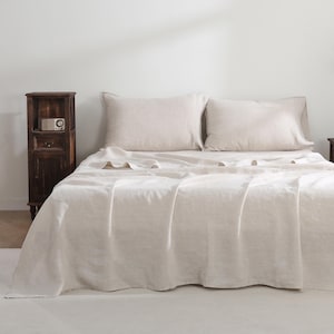 May include: A white linen bed sheet with two white linen pillowcases. The bed is made and the sheet is smooth. The bed is in a bedroom with a wooden nightstand and a white wall.