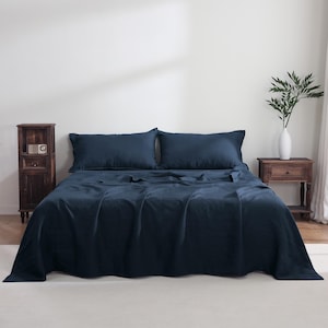 May include: A navy blue bed set, including a fitted sheet, flat sheet, and two pillowcases, is displayed on a bed. The bedding is made of a soft, wrinkle-resistant fabric. A wooden nightstand and a vase with greenery are visible.