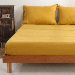 Puede incluir: Una sábana bajera de color amarillo mostaza con un marco de cama de madera. La sábana está hecha de una tela suave y lisa.