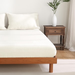 May include: A white bed with a wooden frame and two white pillows. A wooden nightstand with a drawer and a shelf is to the right of the bed. A vase with green leaves is on the nightstand.