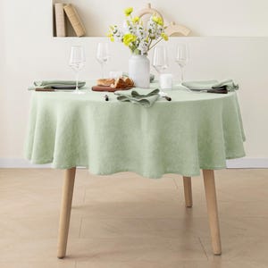 May include: A light green round tablecloth with a subtle texture, set on a table with four place settings, including glasses, silverware, and napkins. A bouquet of yellow and white flowers in a white vase is the centerpiece.