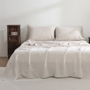 Puede incluir: Un cubrecama de lino blanco con dos almohadas de lino blanco. El cubrecama est&aacute; sobre una cama con una cabecera blanca y una mesita de noche de madera con una peque&ntilde;a radio encima.