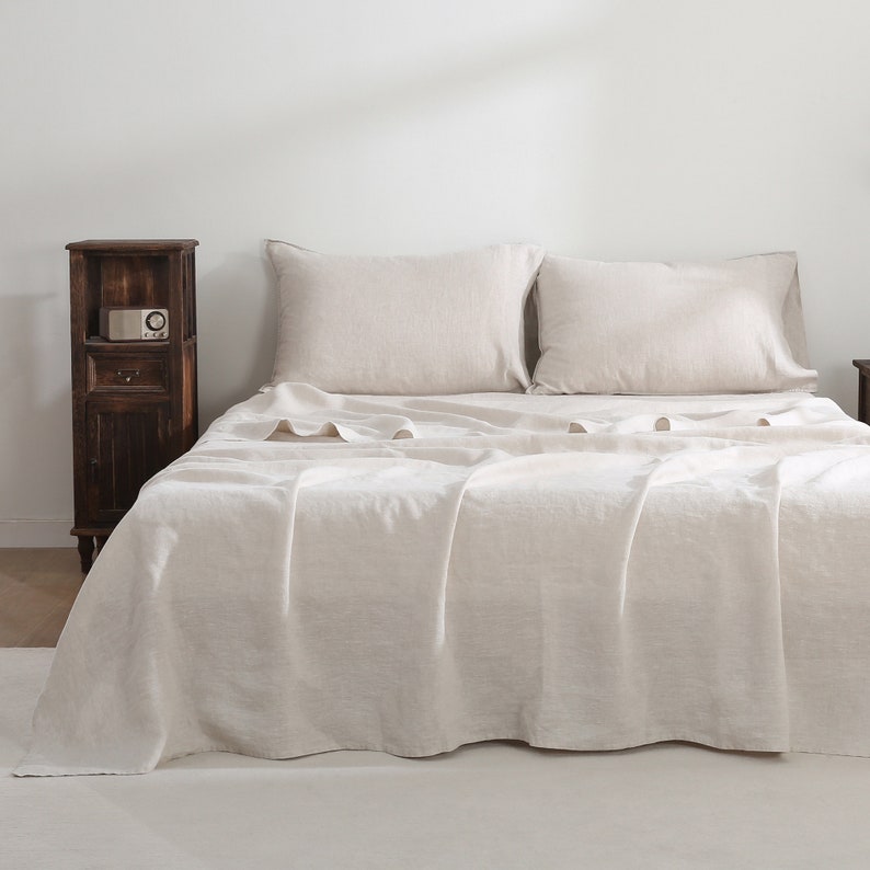 Puede incluir: Un cubrecama de lino blanco con dos almohadas de lino blanco. El cubrecama est&aacute; sobre una cama con una cabecera blanca y una mesita de noche de madera con una peque&ntilde;a radio encima.