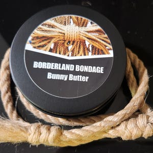 May include: A black tin container with a label that reads "Borderland Bondage Bunny Butter". The container is surrounded by a thick, natural-colored rope.