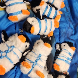 May include: A collection of plush penguin keychains. Each penguin has a black head and back, white body, and orange feet. They are adorned with blue string and small metal keychains. The penguins are arranged on a blue patterned surface.