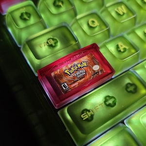 May include: A mechanical keyboard featuring translucent lime green keycaps with gold lettering. A custom red keycap shaped like a Nintendo Game Boy Advance cartridge is installed, displaying the Pokémon FireRed Version game label.