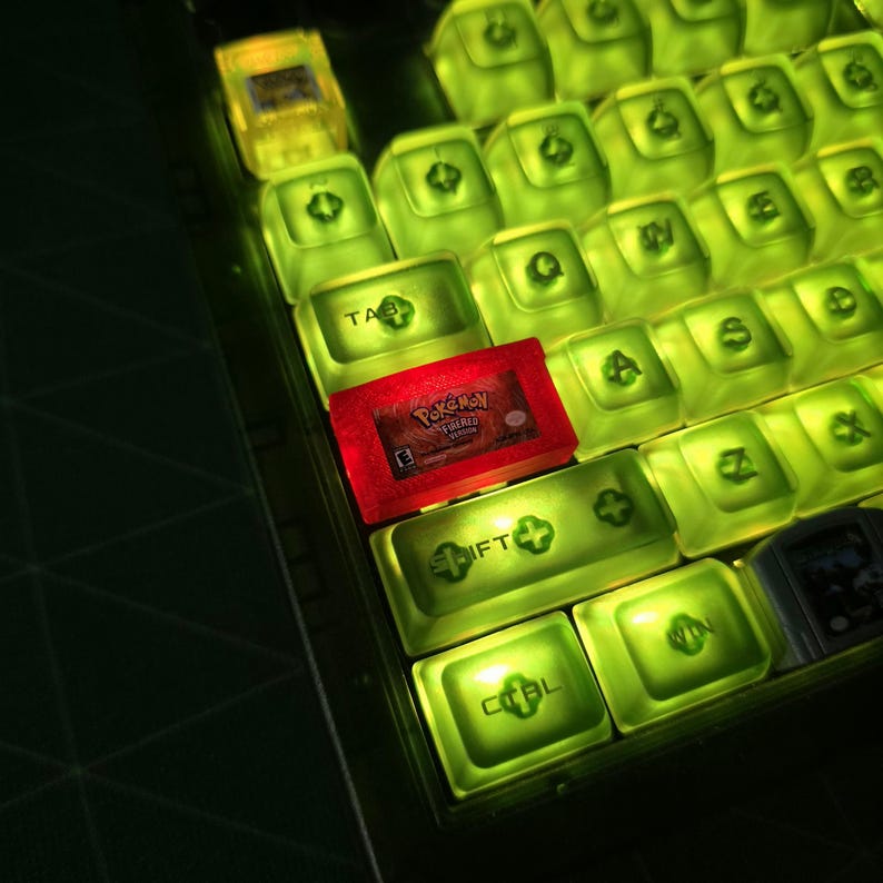 May include: A mechanical keyboard with translucent lime green backlit keycaps and a red translucent Game Boy Advance video game cartridge labeled Pok&eacute;mon FireRed Version resting on the left side of the key layout among the keys.